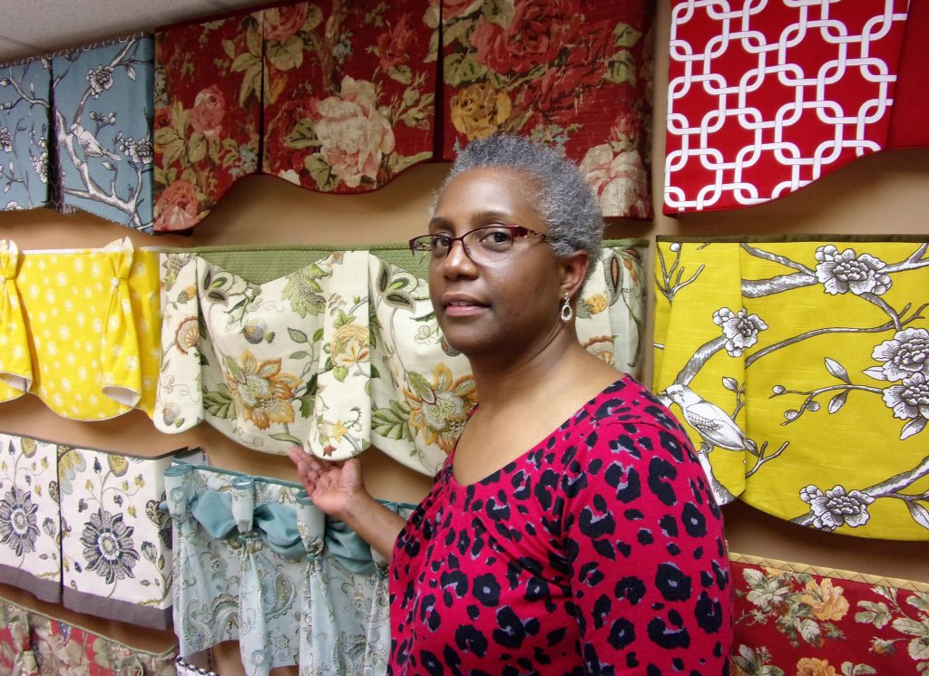 Doshie Witcher, the owner of Black Belt Home Decor, standing in front of a display wall of her Hidden Rod Pocket valance styles.