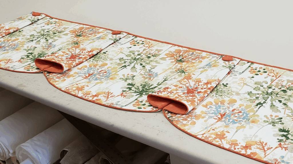 workroom table photo of a custom ANNA Hidden Rod Pocket valance with orange covered buttons at the top of each horn